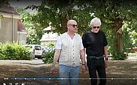Zwei Männer spazieren nebeneinander in einem parkähnlichen Bereich; einer trägt eine gestreifte Weste, der andere ein dunkles Shirt und Sonnenbrille.